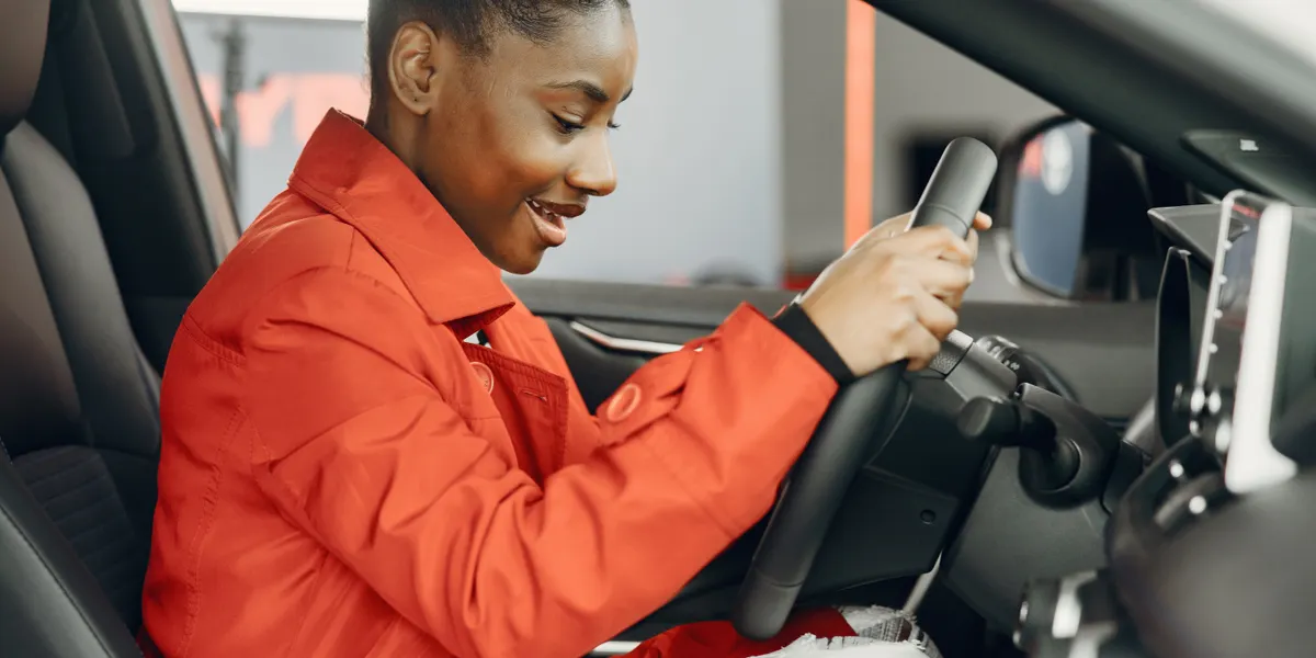 A woman in a red jacket sitting in the driver's seat with one hand on the steering wheel, looking down toward the centre console