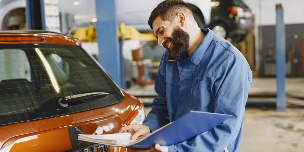 A smiling mechanic in blue overalls reading a service checklist on a clipboard beside the front of an orange hatchback in a workshop